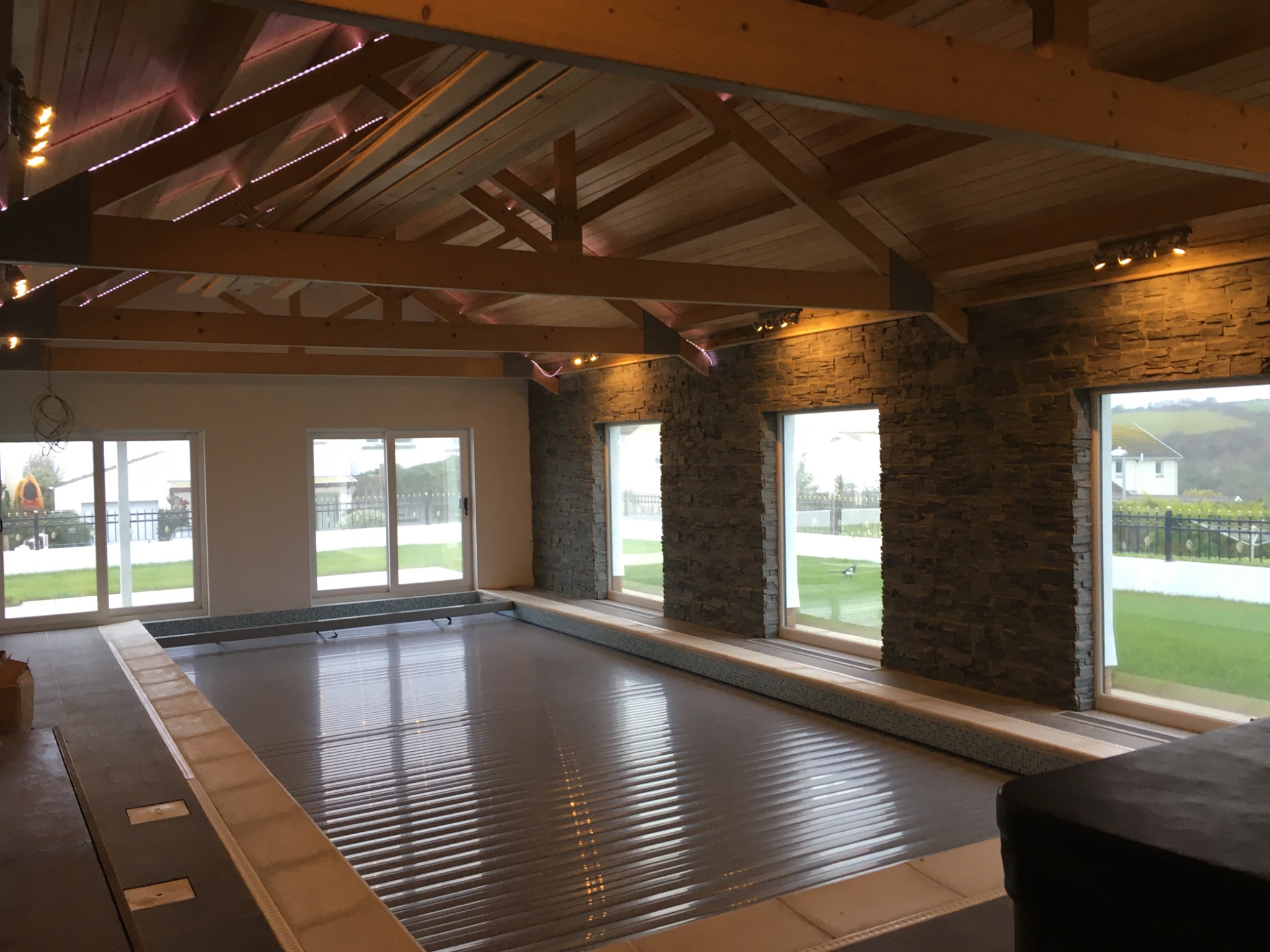 Wnętrze budynku z zadaszonym basenem, automatyczna roleta częściowo zwinięta, ściany wyłożone kamieniem dekoracyjnym, drewniany strop z oświetleniem punktowym