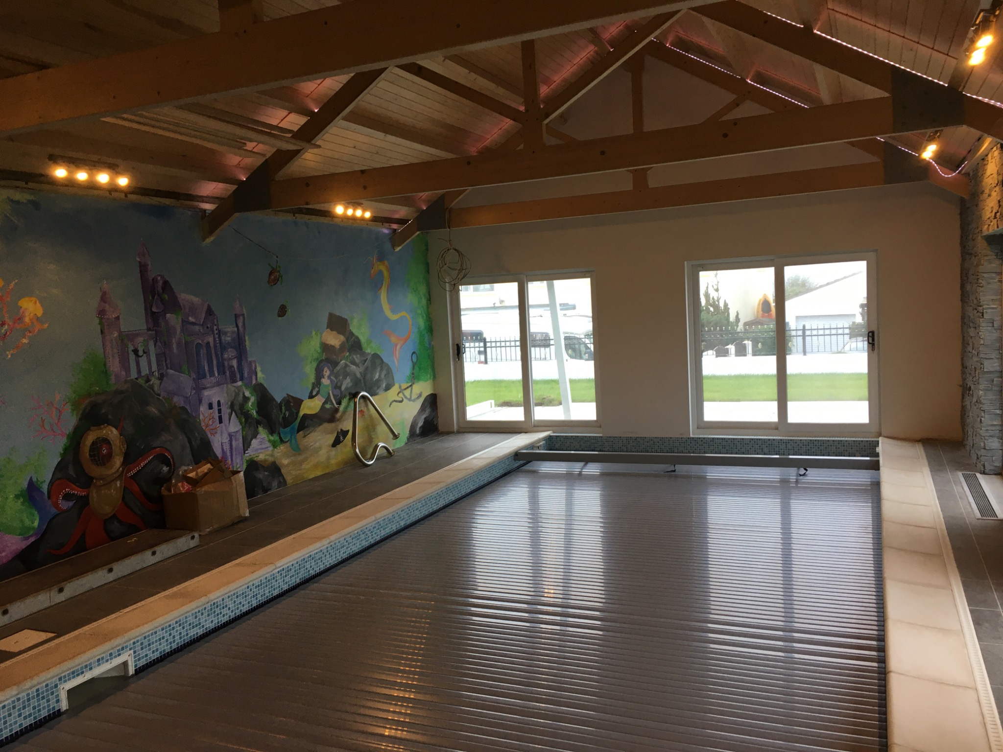 Kryty basen z automatyczną roletą, dekoracyjna ściana z malowidłami podwodnego świata, drewniana konstrukcja dachu z oświetleniem punktowym.