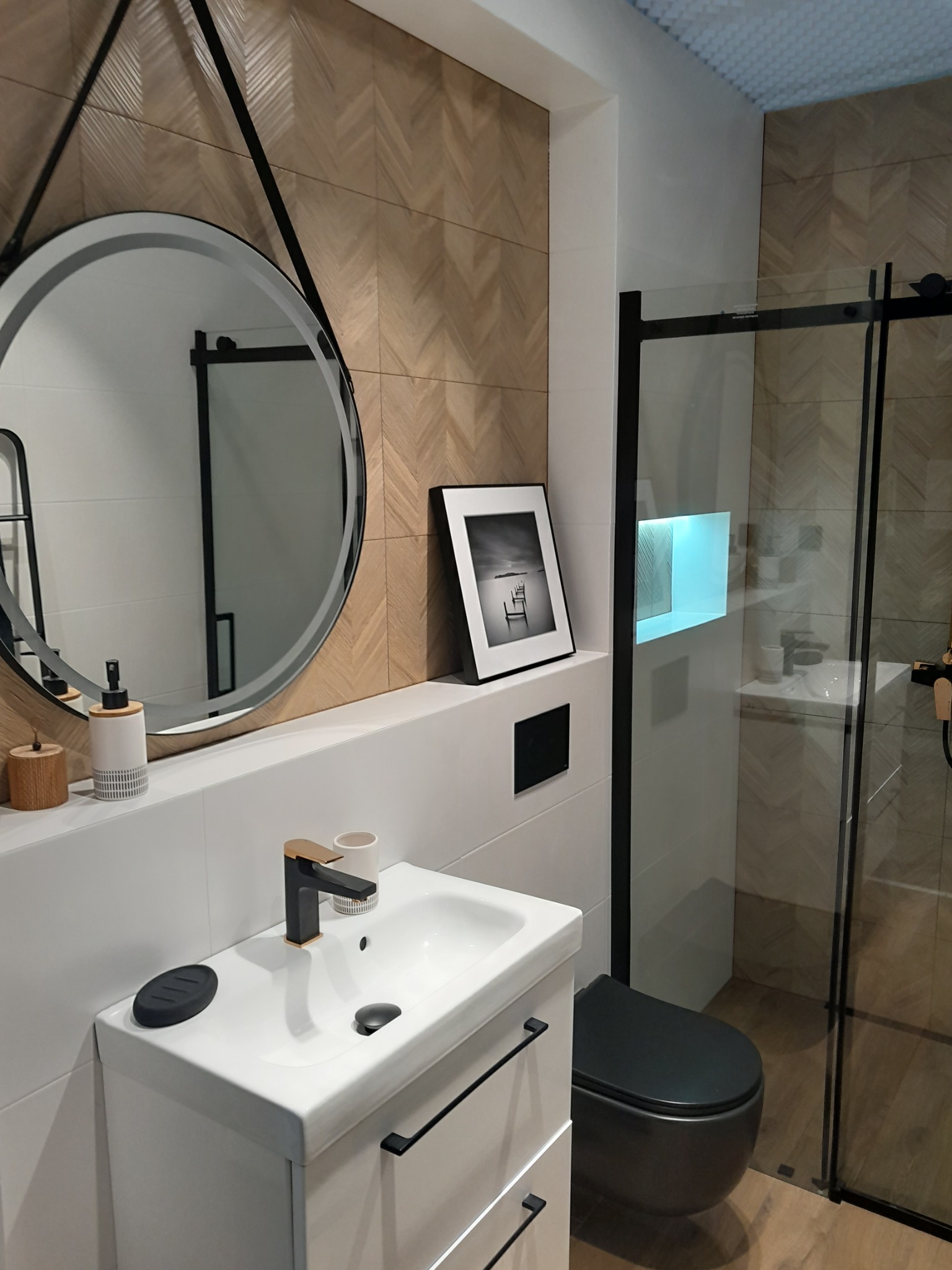 Nowoczesna łazienka z okrągłym lustrem na pasku, umywalką nablatową z czarną armaturą, kabiną prysznicową typu walk-in i toaletą w kolorze antracytu, wykończona płytkami imitującymi drewno...