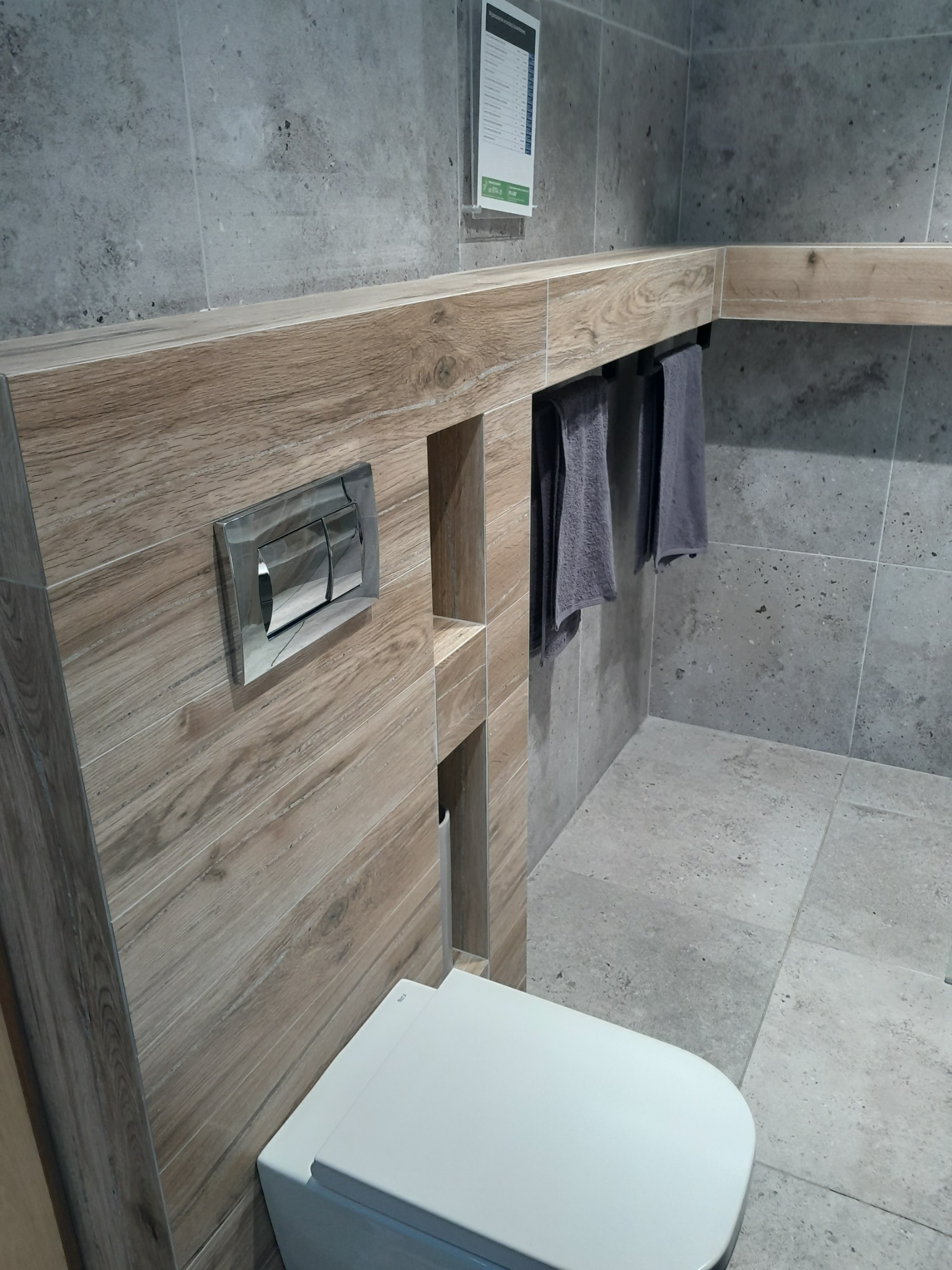 Nowoczesna łazienka z szarymi płytkami imitującymi kamień, drewnopodobną zabudową geberitu z półkami i chromowanym przyciskiem spłukującym oraz wiszącymi szarymi ręcznikami.