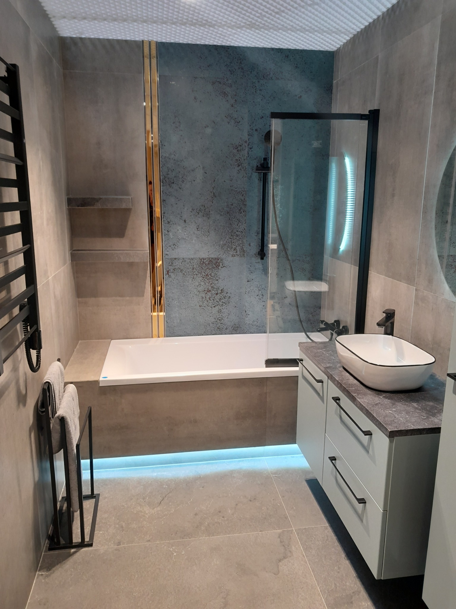 Nowoczesna łazienka z szarymi płytkami, podświetlaną wanną, czarną armaturą i umywalką nablatową o owalnym kształcie.