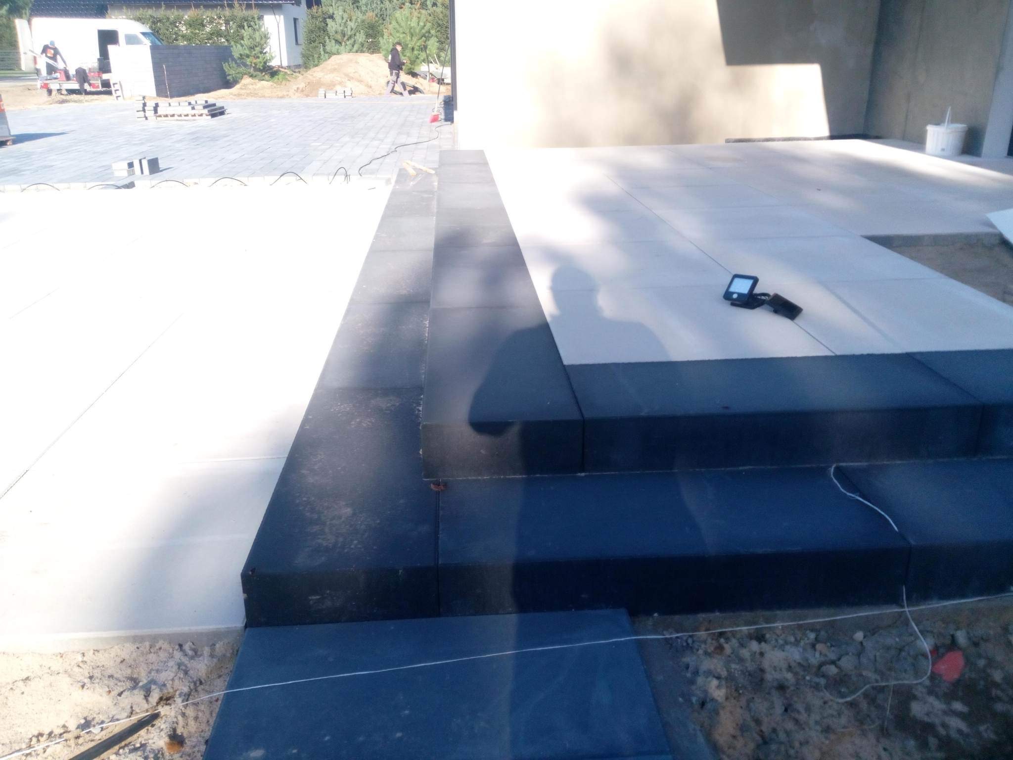 Montaż stopni schodowych z ciemnej kostki brukowej, widoczne sznurki wyznaczające linię, w tle ułożona jasna nawierzchnia, pracownik i samochód dostawczy.