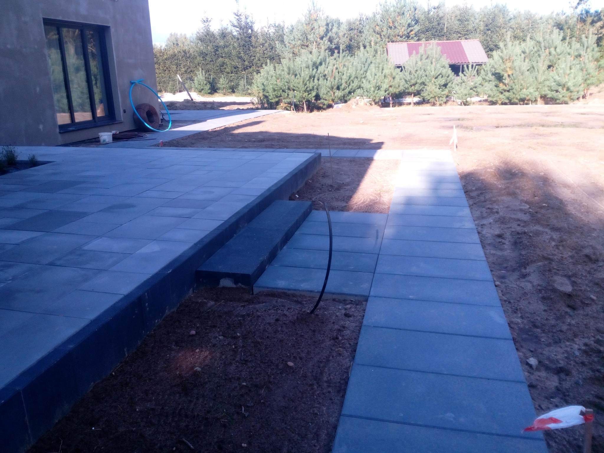 Ułożona, szara kostka brukowa tworząca taras i ścieżkę wokół domu, widok na ogród z młodymi drzewami iglastymi i małą altaną w tle, niebieski wąż ogrodowy oparty o ścianę budynku.