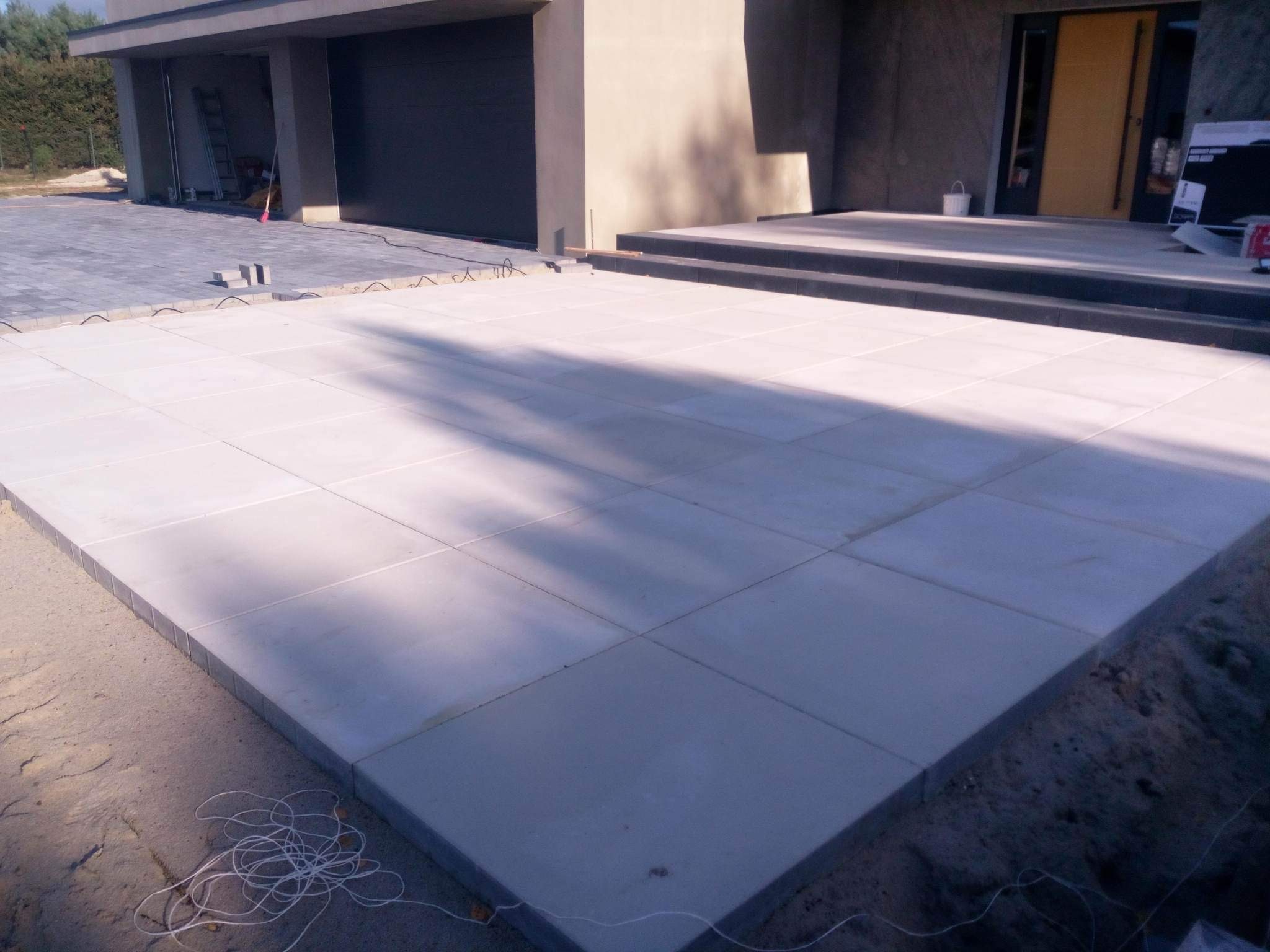 Ułożona jasnoszara kostka brukowa na podjeździe przed nowoczesnym domem z garażem, widoczne przewody pomiarowe na piasku.