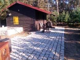 Ułożona kostka brukowa w ogrodzie z drewnianym budynkiem w tle, widoczne drzewa i krzewy, słoneczny dzień.