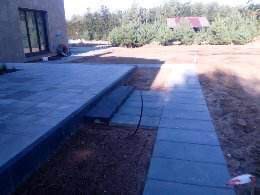 Nowo ułożona kostka brukowa wzdłuż domu, z widocznymi schodami i ścieżką, otoczona przygotowanym terenem pod trawnik, w tle drzewa iglaste.