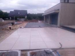 Jasne płyty chodnikowe ułożone na podłożu z piasku, widoczny fragment domu z szarą elewacją i przygotowany teren pod dalsze prace.