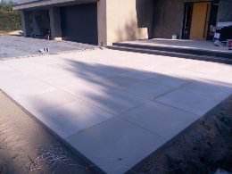 Jasnoszara, prostokątna nawierzchnia z płyt betonowych przed budynkiem z ciemnymi elementami architektonicznymi, widoczny fragment podjazdu z szutrem.