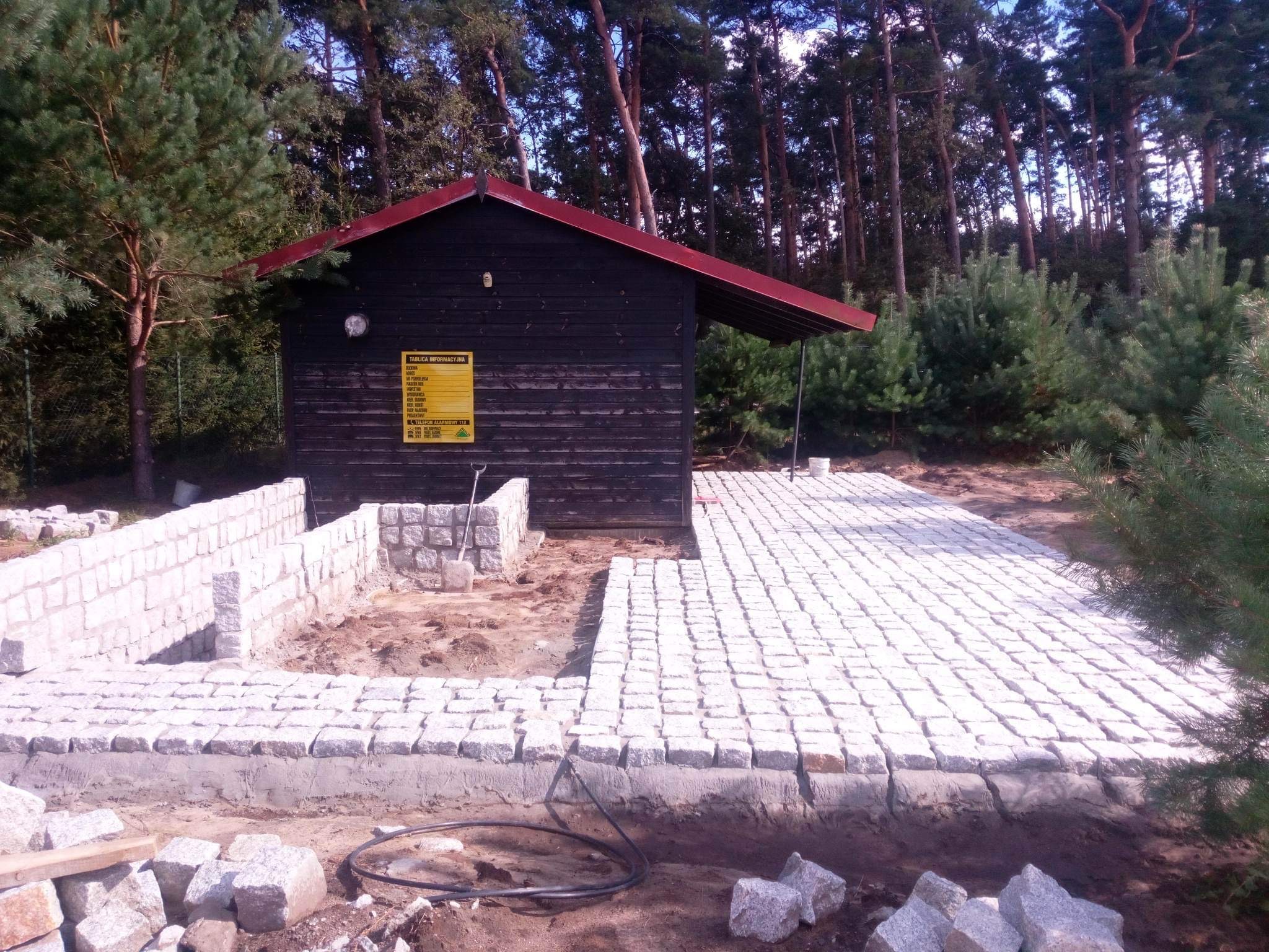 Fragment posesji w trakcie budowy z ułożoną częścią nawierzchni z kostki granitowej, widoczne fundamenty pod murek z kamienia, czarny drewniany budynek gospodarczy z żółtą tablicą informacyjną...