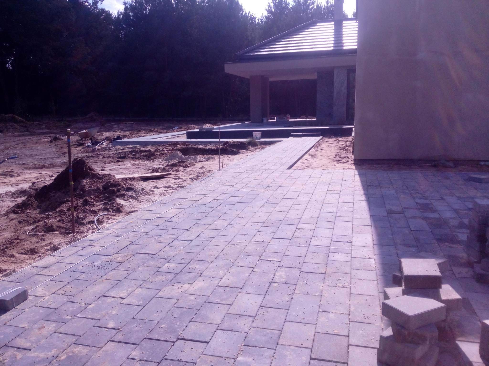 Świeżo ułożona ścieżka z kostki brukowej prowadząca do zadaszonej konstrukcji w trakcie budowy, widoczny stos kostki, teren przygotowany pod dalsze prace.