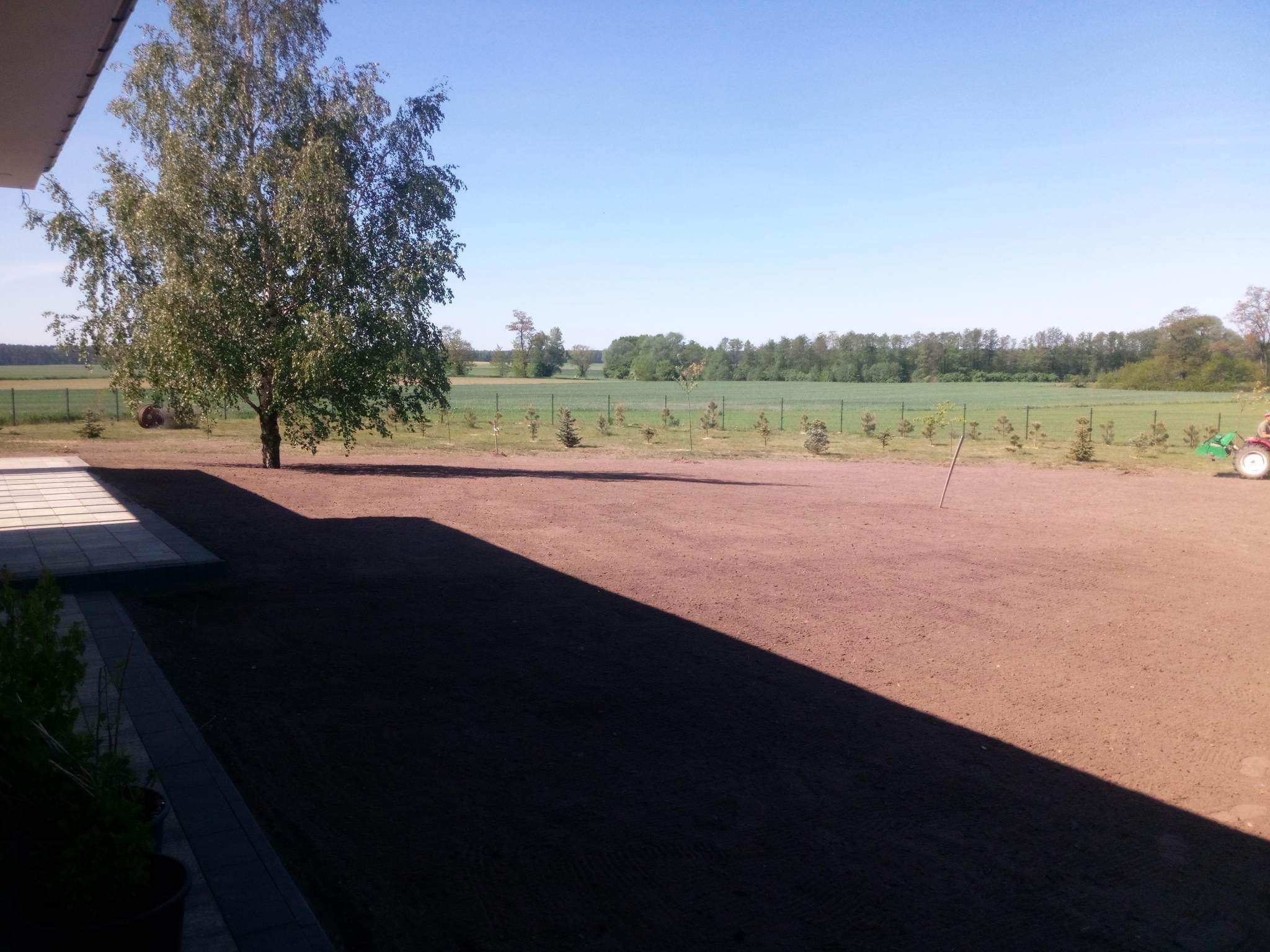 Przygotowany teren pod trawnik z widocznym świeżo nawiezionym humusem, w tle drzewa i zieleń, częściowo zacieniony fragment z kostką brukową i roślinnością.