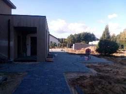 Nowo ułożona, grafitowa kostka brukowa prowadząca do nowoczesnego domu z płaskim dachem. W tle widoczny teren przygotowany pod dalsze prace, ogrodzenie oraz drzewa.