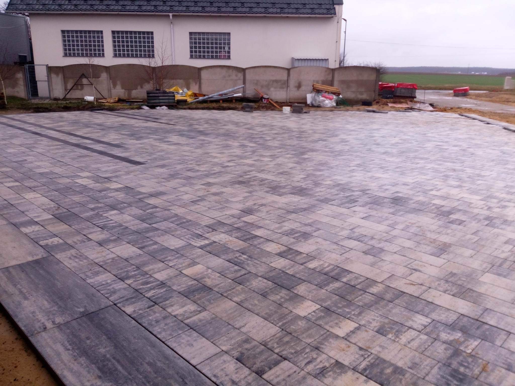 Ułożona kostka brukowa w odcieniach szarości i bieli przed budynkiem z szarym dachem, widoczne materiały budowlane, betonowe ogrodzenie i fragment pola w tle, pochmurny dzień.