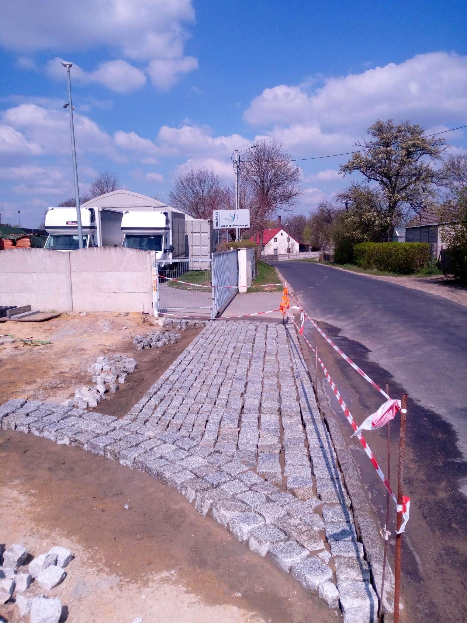 Fragment układanej ścieżki z kostki granitowej, widoczne pakiety kostki do ułożenia, w tle brama, budynki magazynowe i droga asfaltowa.