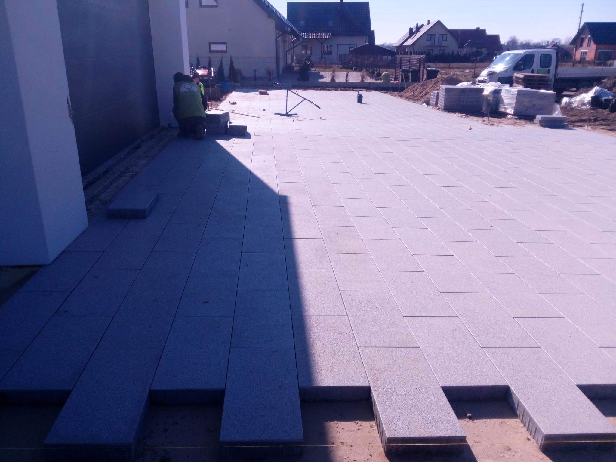 Układanie prostokątnej kostki brukowej w jasnoszarym odcieniu na podjeździe, widoczni pracownicy w odblaskowych kamizelkach, częściowo ułożona nawierzchnia, w tle domy jednorodzinne i samochód...