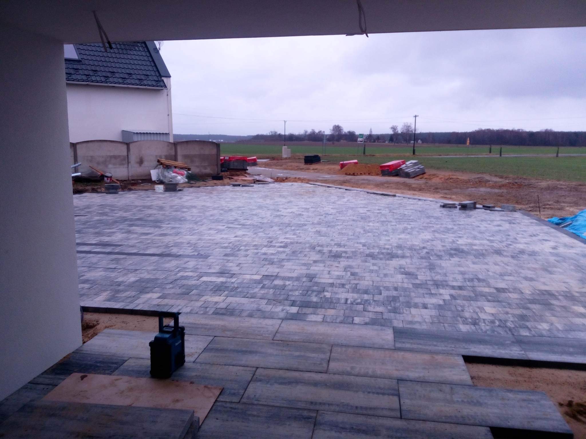Szara kostka brukowa ułożona na podjeździe przed domem w trakcie prac wykończeniowych, widoczne palety z kostką w tle i narzędzie budowlane w rogu.
