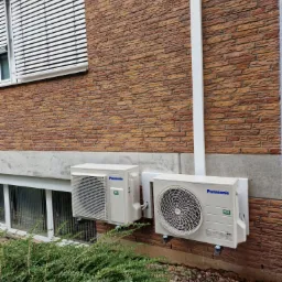 Dwa klimatyzatory Panasonic zamontowane na ścianie z cegły, widoczne przewody i rury, okna z żaluzjami w tle, krzewy i kamienie na pierwszym planie.