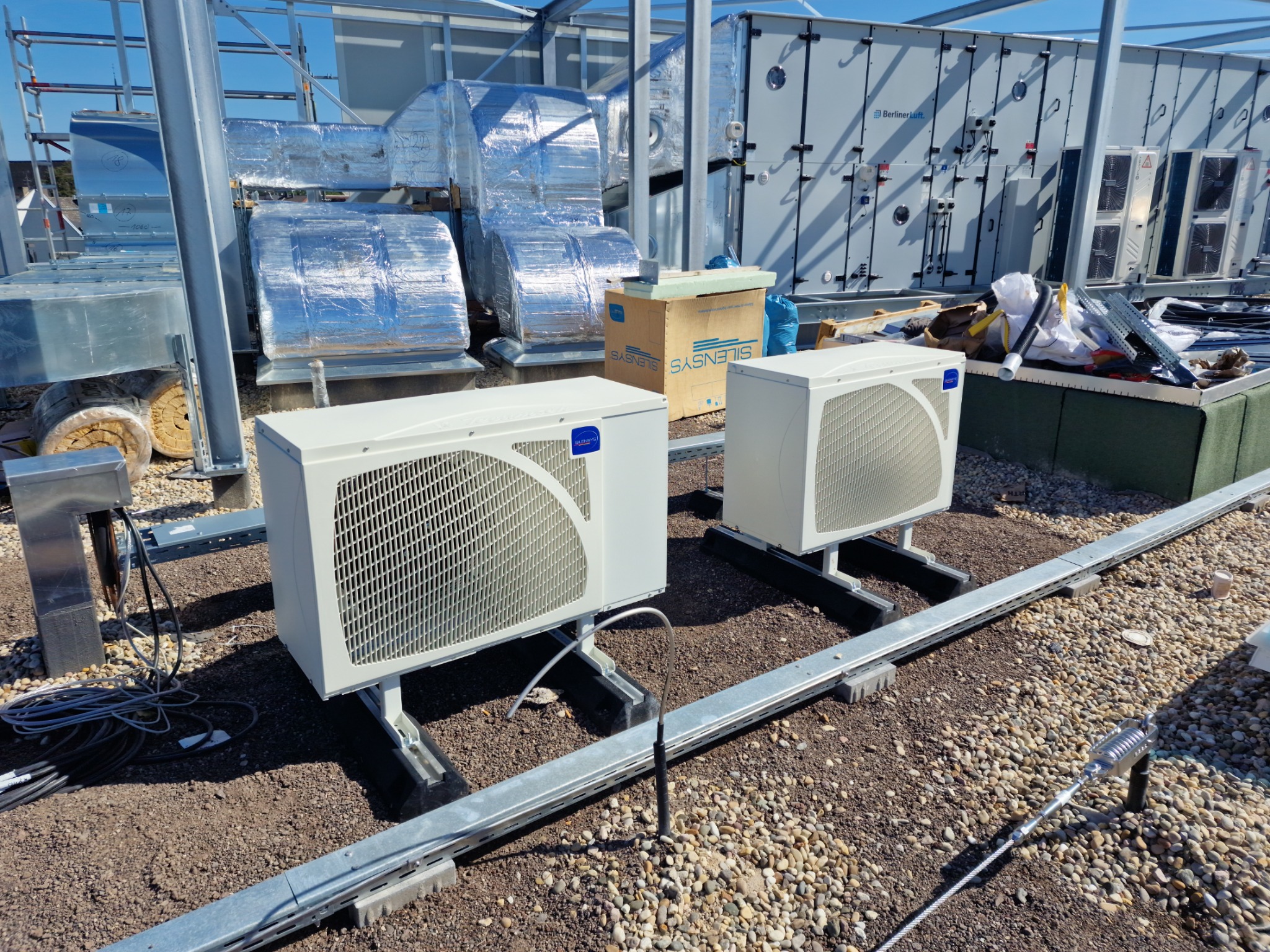 Dwa białe agregaty klimatyzacyjne na dachu budynku, widoczne elementy instalacji, w tle srebrne kanały wentylacyjne owinięte folią izolacyjną i centrala wentylacyjna BerlinerLuft.