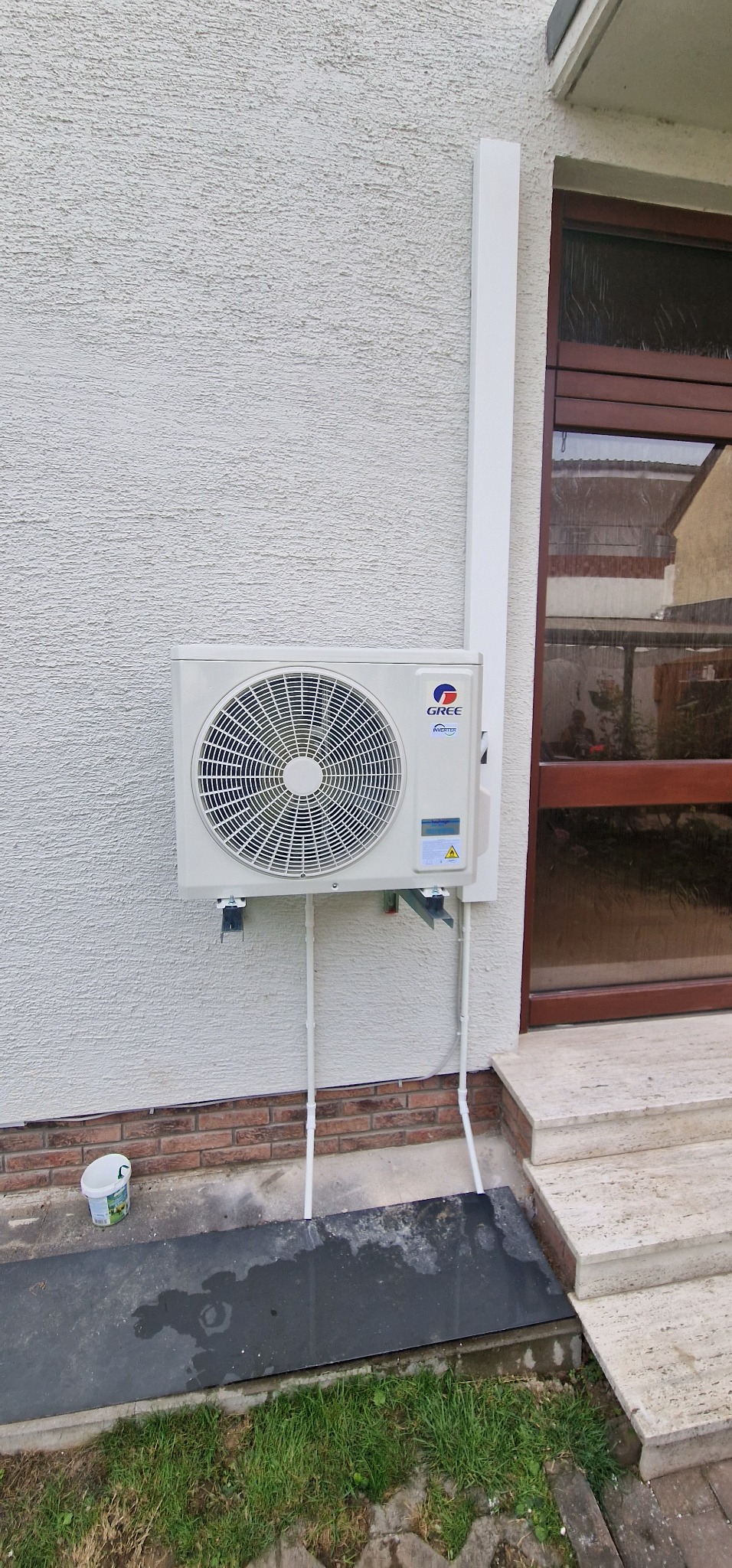 Zewnętrzna jednostka klimatyzacji Gree Inverter zamontowana na elewacji domu z widocznymi rurami odprowadzającymi skropliny i puszką po farbie obok.