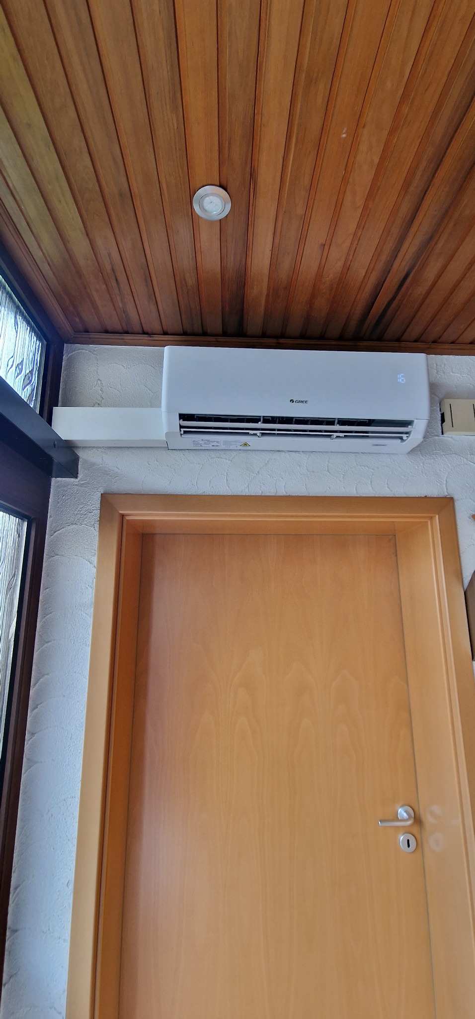 Biały klimatyzator marki Gree zamontowany nad drewnianymi drzwiami w pomieszczeniu z drewnianym sufitem i oknem po lewej stronie.