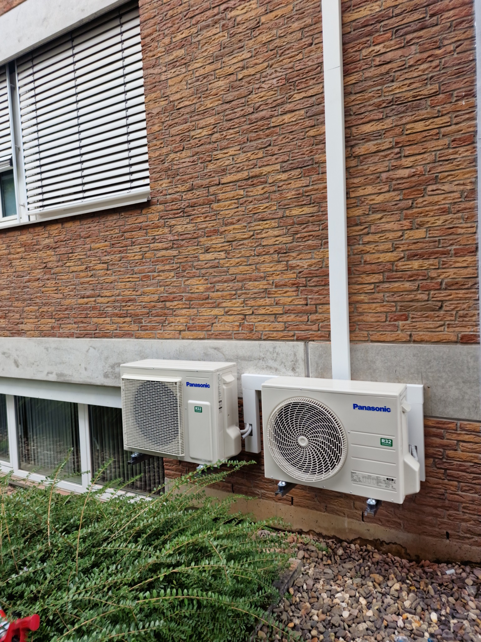 Dwa klimatyzatory Panasonic zamontowane na ścianie z cegły, widoczne przewody i rury, okna z żaluzjami w tle, krzewy i kamienie na pierwszym planie.