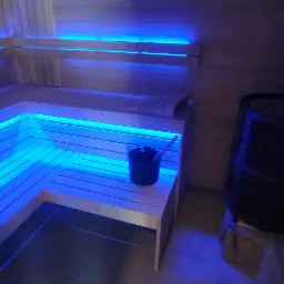 Wnętrze sauny z drewnianymi ławkami podświetlonymi niebieskim światłem LED, piec w rogu i wiaderko z chochlą na ławce.