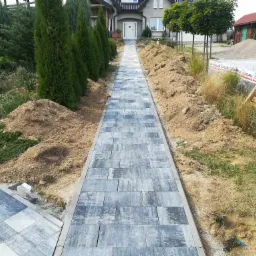 Długa, nowo ułożona ścieżka z szarej kostki brukowej prowadząca do białych drzwi domu, obrzeża obsypane ziemią, w tle widoczne drzewa i krzewy.