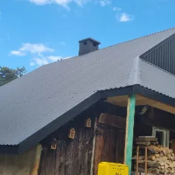 Ciemnoszary, blaszany dach z kominem na drewnianym domu z bali, widoczne drewno na opał i skrzynka gazowa na zewnątrz.