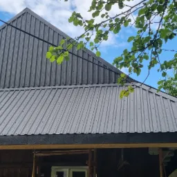 Nowo pokryty blachą trapezową dach domu z widocznymi elementami konstrukcyjnymi, na tle błękitnego nieba i zieleni drzew.