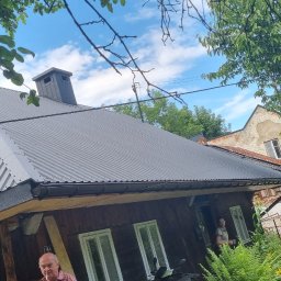 DOMY KONRAD MAJKUT - Ukończony remont dachu z blachy falistej na drewnianym domu, widoczny komin i fragment sąsiedniego budynku.