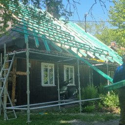 DOMY KONRAD MAJKUT - Robotnik w kraciastej koszuli niesie zielone belki na tle starego, drewnianego domu w trakcie remontu dachu, otoczonego rusztowaniem i stosem drewna.