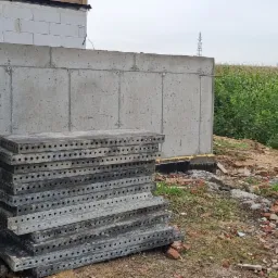 Betonowy fundament domu w trakcie budowy, z widocznymi śladami po szalunkach i stosem metalowych płyt szalunkowych obok, w tle pole kukurydzy.