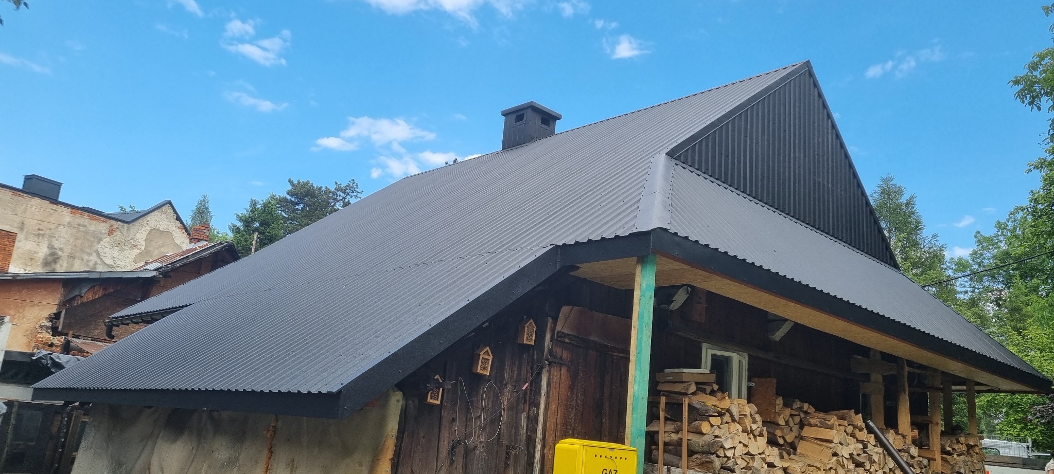 Ciemnoszary, blaszany dach z kominem na drewnianym domu z bali, widoczne drewno na opał i skrzynka gazowa na zewnątrz.