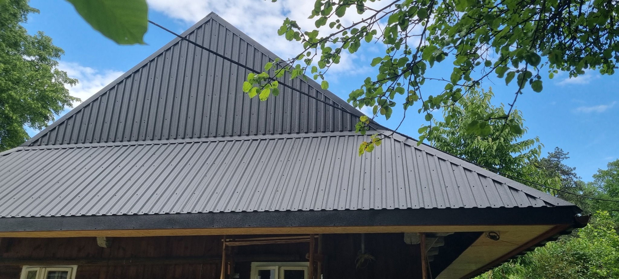 Nowo pokryty blachą trapezową dach domu z widocznymi elementami konstrukcyjnymi, na tle błękitnego nieba i zieleni drzew.