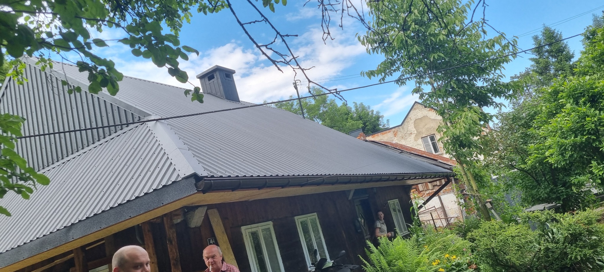 Ukończony remont dachu z blachy falistej na drewnianym domu, widoczny komin i fragment sąsiedniego budynku.