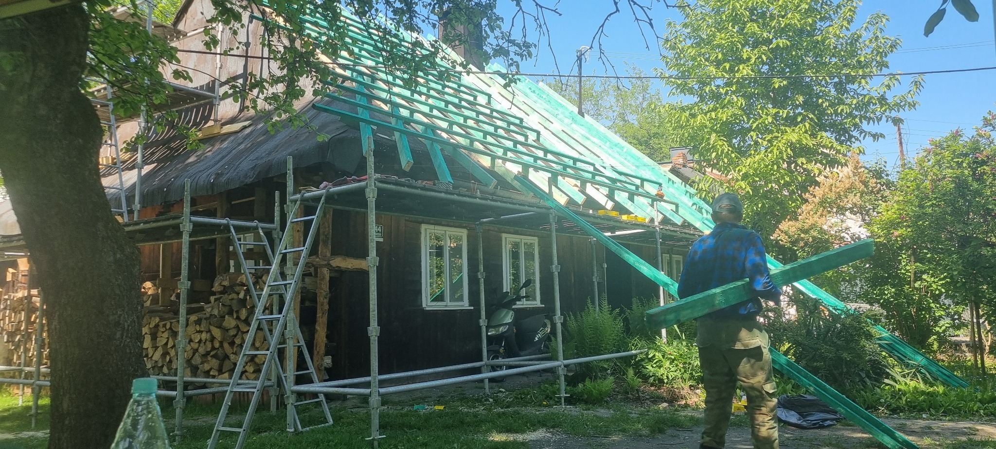 Robotnik w kraciastej koszuli niesie zielone belki na tle starego, drewnianego domu w trakcie remontu dachu, otoczonego rusztowaniem i stosem drewna.