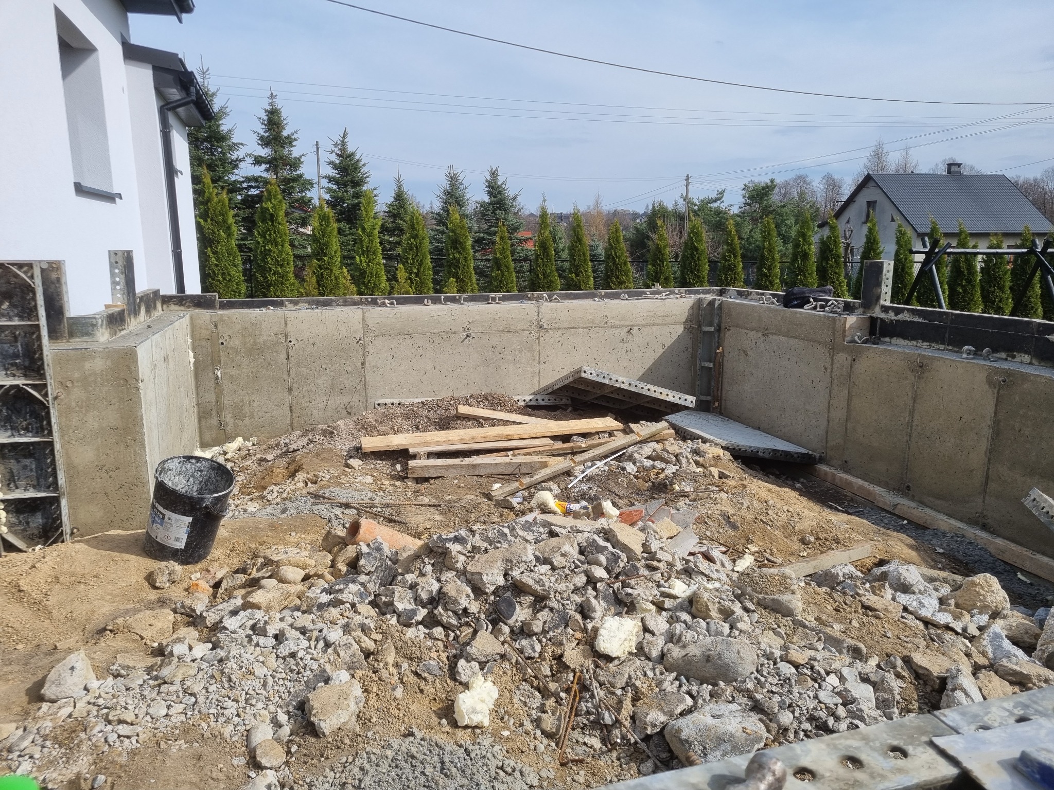 Wykop pod fundamenty domu jednorodzinnego z widocznymi betonowymi ścianami szalunkowymi, gruzem i drewnianymi elementami szalunku, w tle rząd iglaków i budynek mieszkalny.