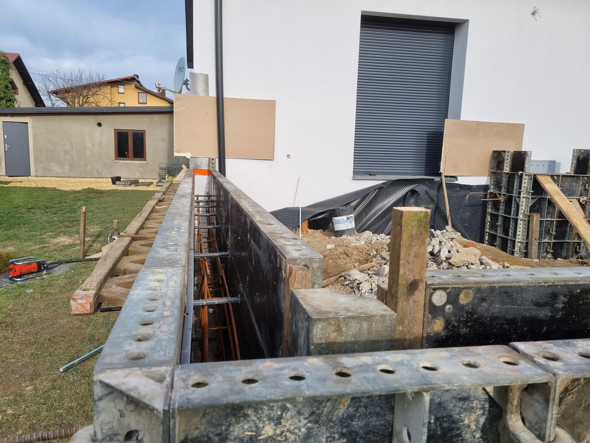 Szczegółowe ujęcie szalunku fundamentowego z widocznymi prętami zbrojeniowymi, w tle biały budynek z szarą roletą i fragment sąsiedniego domu, na ziemi gruz i materiały budowlane.