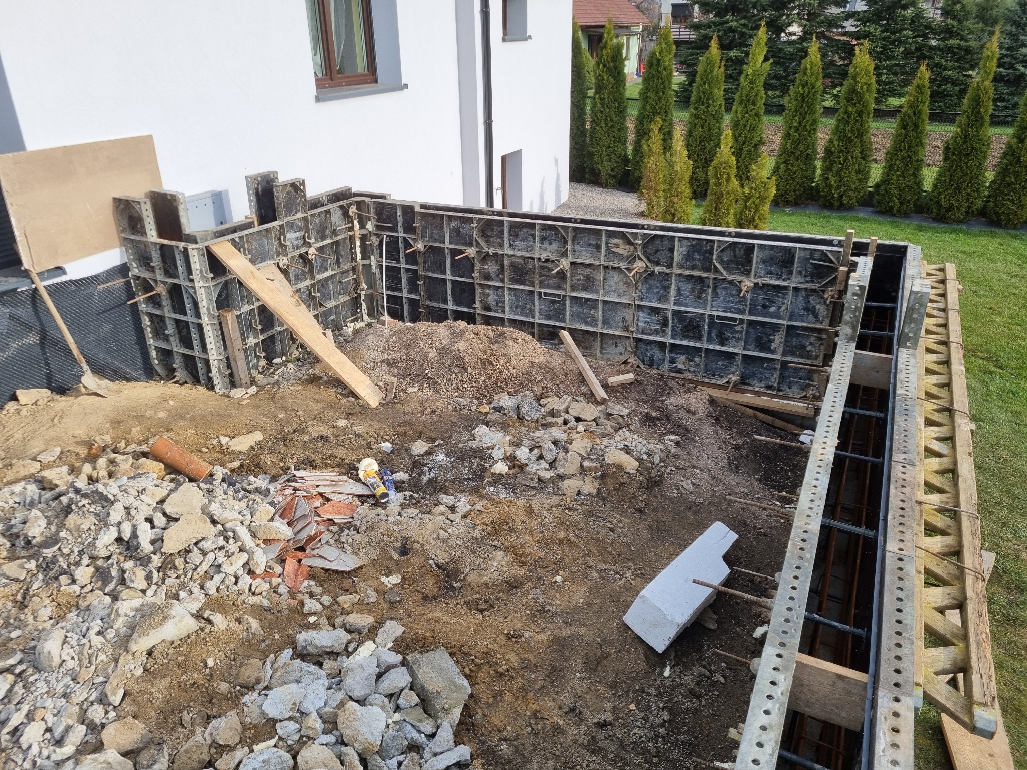 Wykop pod fundamenty domu jednorodzinnego z widocznym szalunkiem i zbrojeniem, gruzem oraz elementami konstrukcyjnymi na pierwszym planie, w tle biała ściana budynku i rząd iglaków.