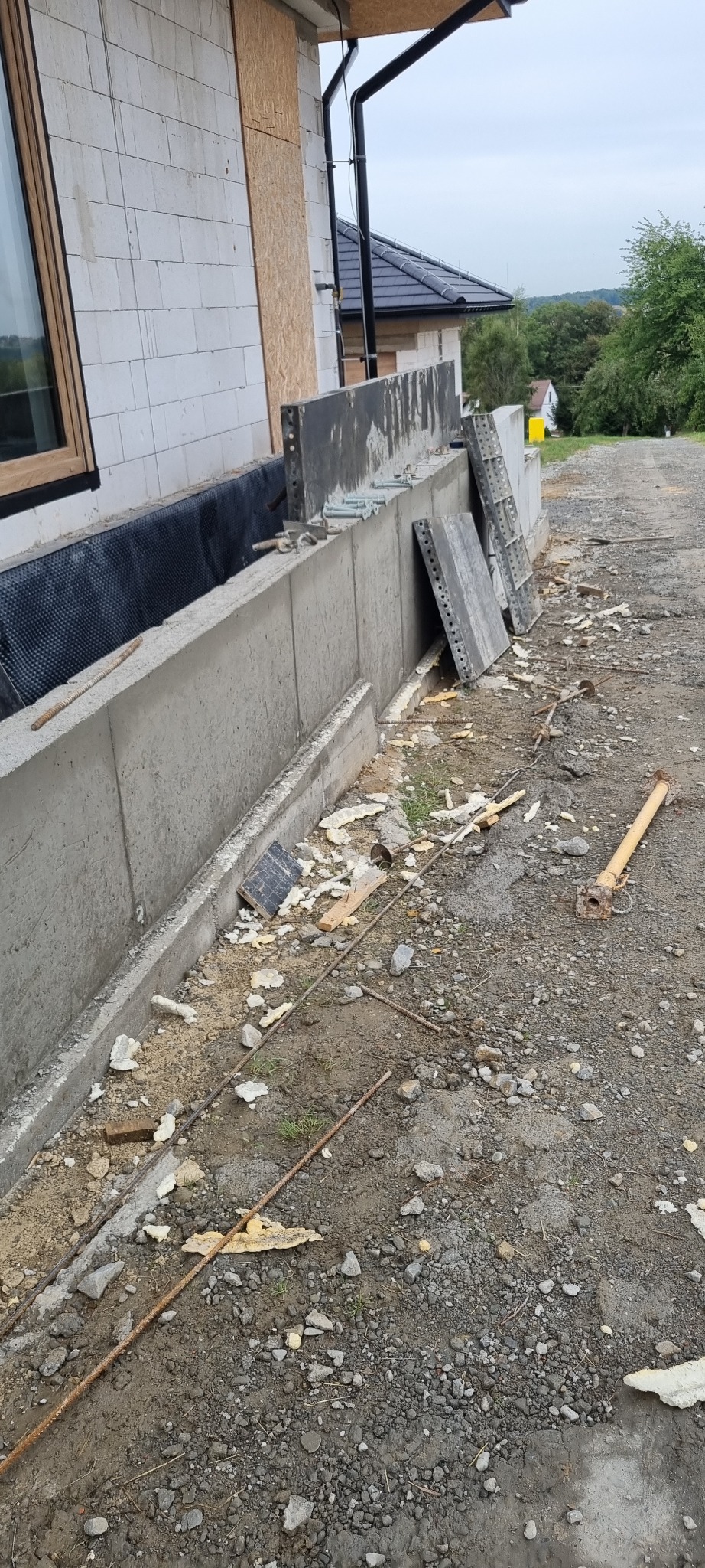 Wylany fundament z hydroizolacją, widoczne szalunki i wystające pręty zbrojeniowe, elementy drewniane i gruz na ziemi obok budynku w trakcie budowy.