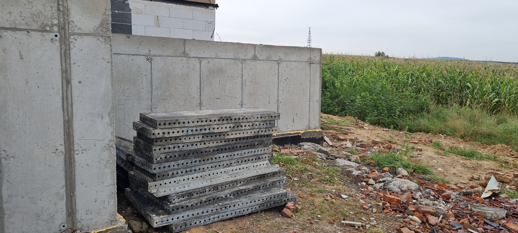 Betonowy fundament domu w trakcie budowy, z widocznymi śladami po szalunkach i stosem metalowych płyt szalunkowych obok, w tle pole kukurydzy.