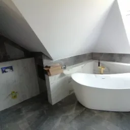 Nowoczesna wanna wolnostojąca w łazience na poddaszu z marmurowymi płytkami i złotą armaturą, kartonowe opakowania po akcesoriach jeszcze nie usunięte.