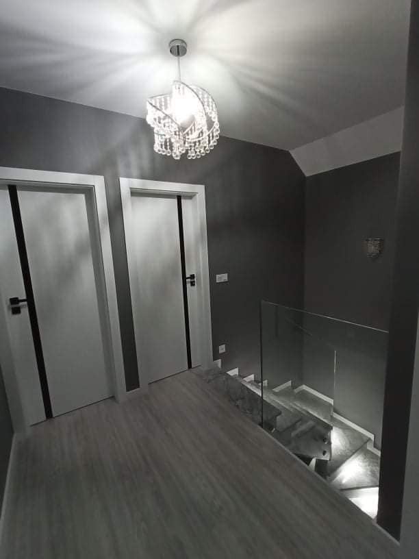 Szare wnętrze z drewnopodobną podłogą, białymi drzwiami z czarnymi akcentami, szklaną balustradą schodów i kryształowym żyrandolem.