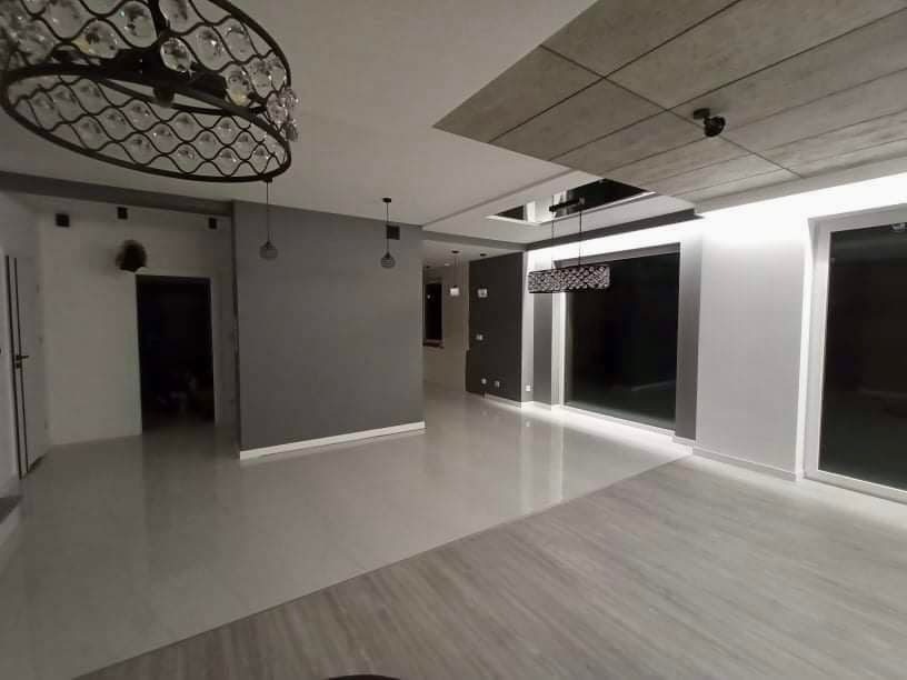 Wnętrze przestronnego, nowocześnie wykończonego salonu z błyszczącą podłogą, szarą ścianą, ozdobnym żyrandolem i designerskim sufitem z oświetleniem punktowym.