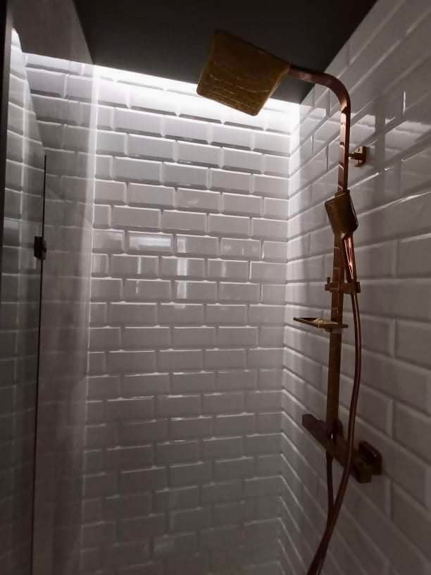 Nowoczesna kabina prysznicowa z białymi, cegiełkowymi płytkami i miedzianą armaturą, w tym deszczownicą i słuchawką.