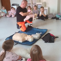 Rafał Radecki - Instruktor w odzieży roboczej demonstruje resuscytację niemowlęcia na manekinie podczas szkolenia z pierwszej pomocy dla dzieci. Grupa dzieci obserwuje.