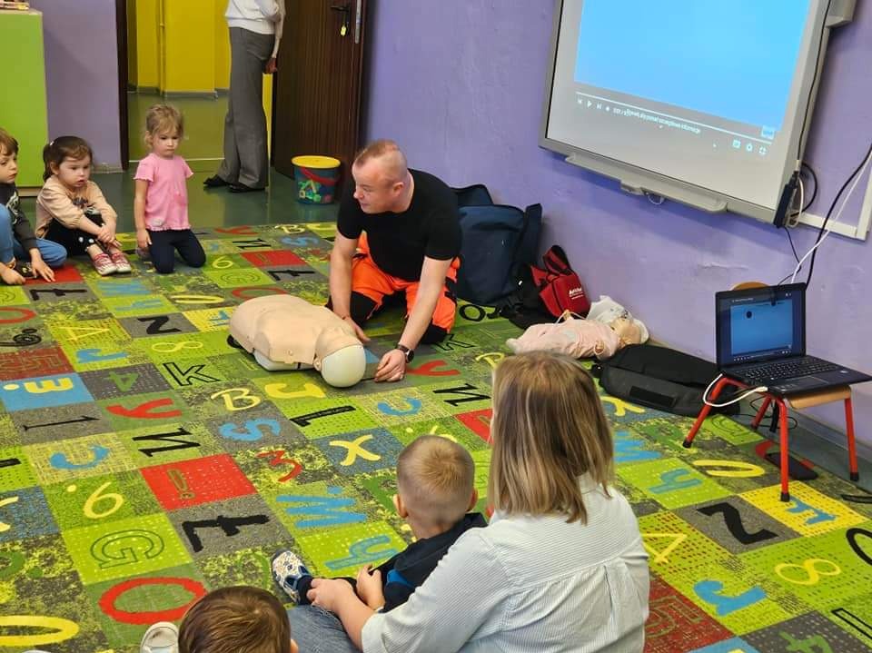 Instruktor w pomarańczowych spodniach demonstruje resuscytację na manekinie przed grupą dzieci na kolorowym dywanie z literami i cyframi. Szkolenie BHP dla najmłodszych.