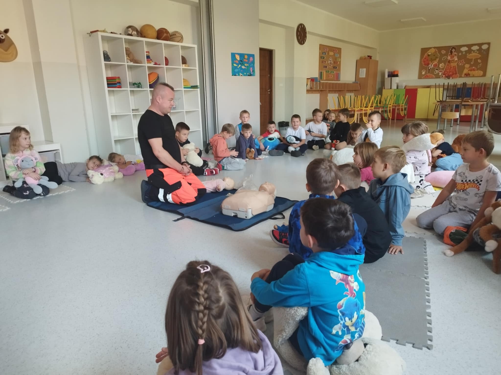 Instruktor demonstruje resuscytację krążeniowo-oddechową na manekinie przed grupą dzieci siedzących na podłodze w przedszkolu. Widoczny sprzęt ratowniczy.