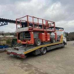 Pomarańczowy podnośnik nożycowy Genie GS-3384, z oznaczeniem 4x4, zabezpieczony pasami, przewożony na platformie lawety z żółtymi elementami, na tle pochmurnego nieba i przemysłowego otoczenia.