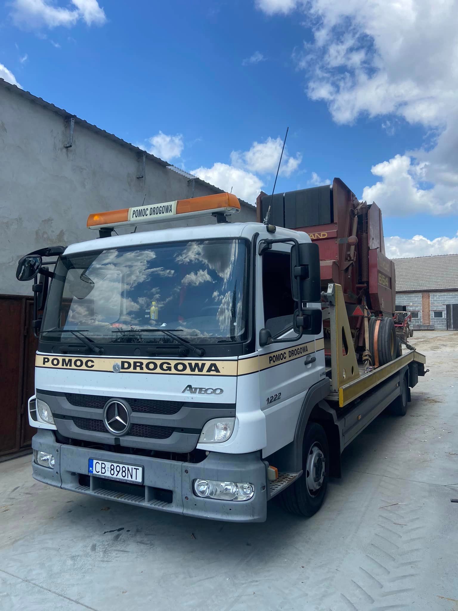 Biała ciężarówka Mercedes Atego z napisem 'Pomoc Drogowa' przewozi na platformie czerwoną maszynę rolniczą, widoczna tablica rejestracyjna CB 898NT, tło stanowią budynki gospodarcze i błękitne...